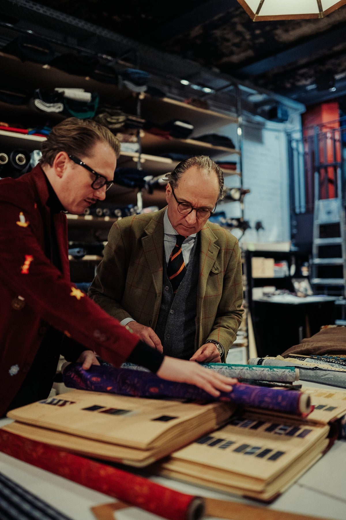 Jan-Henrik M. Scheper-Stuke und ein Herr im Tweed-Sakko prüfen am Zuschnitttisch einen violetten Paisley-Seidenballen, davor aufgeschlagene Musterbücher